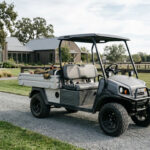 A charcoal gray utility golf cart for farm use featuring an aluminum cargo bed and all-terrain tires, parked on a gravel path of a professional estate.